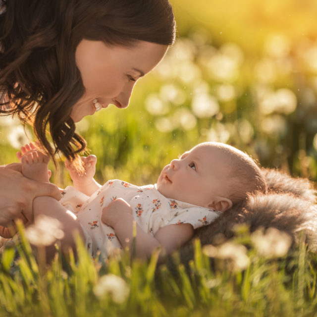 baby laying on grass looking up at her mom and smiling back light flowers - boulder photographer
